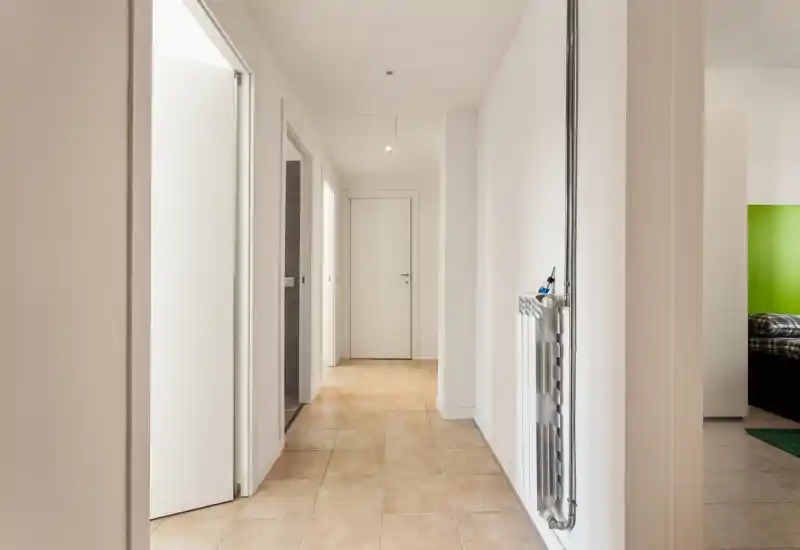 Bright, minimal hallway with tiled floor, white walls and a radiator; clean corridor leading to interior doors, suitable for an apartment entrance or landing.