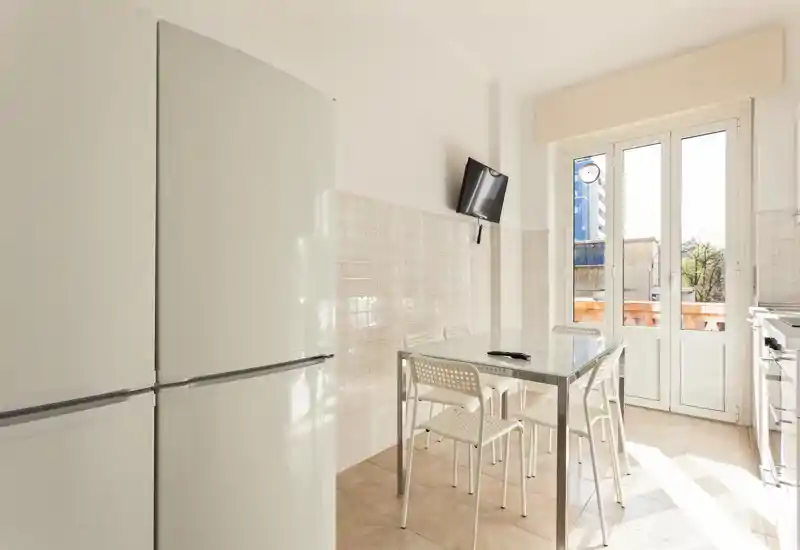 Bright, clean kitchen with a refrigerator, glass-top dining table and four chairs next to a large window/door letting in abundant natural light.