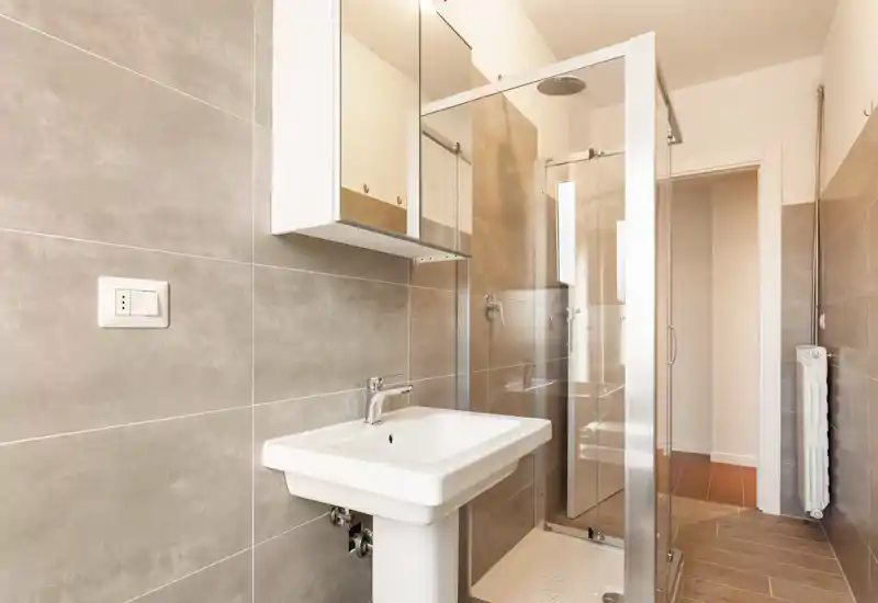 Bright, modern bathroom with a glass shower enclosure, wall-mounted mirror cabinet and pedestal sink; neutral large-format tiles create a clean contemporary look.