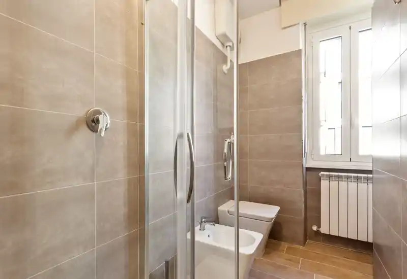 Modern compact bathroom with a glass shower cabin, bidet and toilet, neutral beige tiles and a bright window providing natural light.