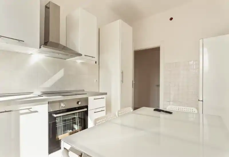 Bright modern kitchen with white cabinets, built-in oven, extractor hood and a glossy dining table with chairs — ideal for small-family meals and light-filled cooking.