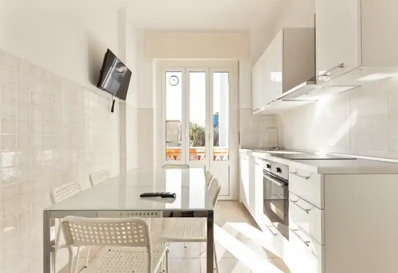 Bright modern kitchen with a glass-topped dining table and simple white cabinetry. Natural light floods the space from a tall door leading to a small balcony, creating a fresh, minimal atmosphere ideal for short-term rentals.