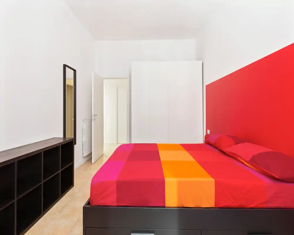 Dormitorio moderno y luminoso con una gran cama doble colorida, mueble de almacenamiento minimalista y un espejo alto. Paredes blancas y cabecero rojo que aportan un estilo contemporáneo.