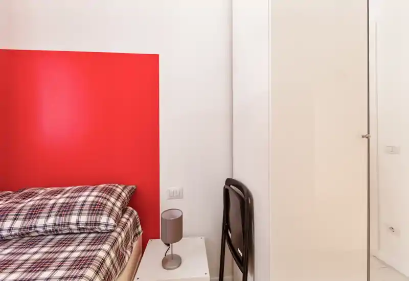 Chambre moderne et compacte avec couette à carreaux, tête de lit rouge en accent et une lampe de chevet ; lumineuse et au mobilier minimaliste.