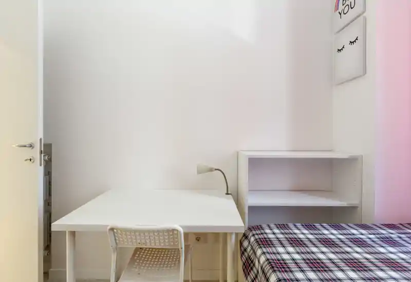 Coin chambre lumineux et minimaliste avec un lit simple, un bureau blanc et une étagère. La composition met en valeur un espace de travail rangé à côté du lit, idéal pour étudiants ou une personne seule.