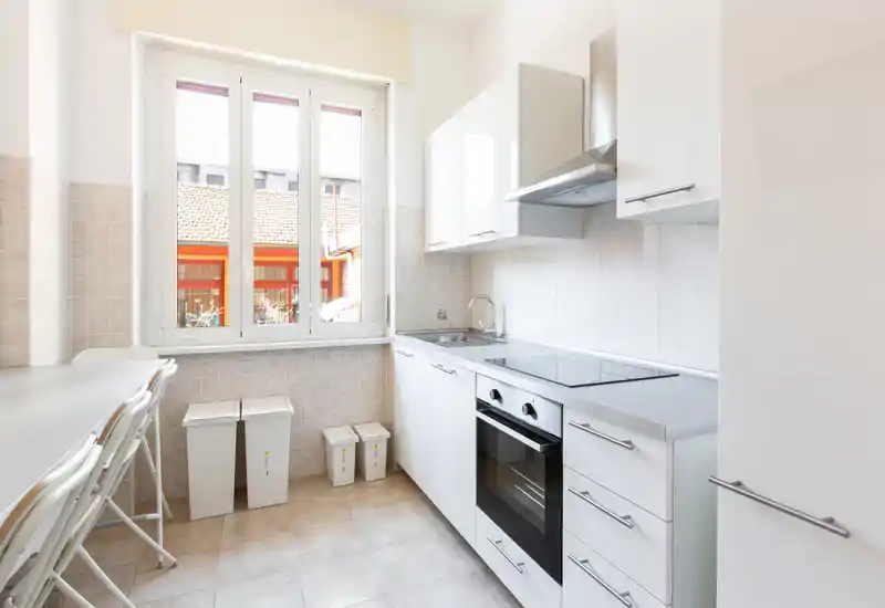 Cuisine moderne et lumineuse avec meubles blancs, four encastré et plaque vitrocéramique, évier sous une grande fenêtre et petit comptoir avec tabourets.