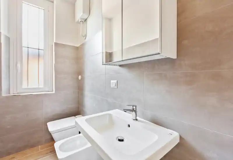 Bright, modern bathroom with wall-mounted mirror cabinet, white sink and bidet/toilet; neutral grey tiles and a tall frosted window provide natural light.