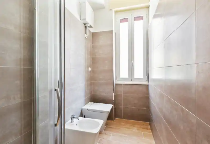 Bright, modern bathroom with tiled walls, a bidet and toilet, glass shower door and a tall window providing natural light.