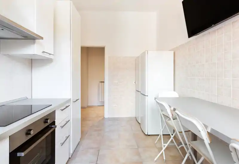 Bright, modern kitchen with integrated oven, induction cooktop, fridge and a small dining counter with folding chairs — clean neutral tiles and a minimal, functional layout.