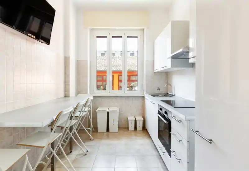 Bright, modern galley kitchen with white cabinets, integrated oven and a long breakfast counter with folding chairs under a large window.