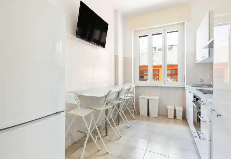 Cucina luminosa e moderna con TV a parete, piano d'appoggio con quattro sedute, frigorifero e forno incassato — ambiente pulito e ben illuminato, ideale per pasti veloci.