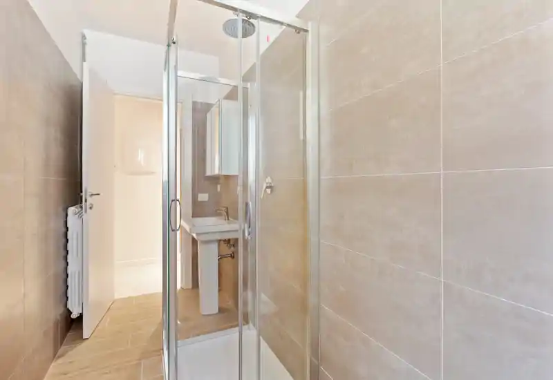 Bright, modern bathroom with a glass-enclosed shower and a pedestal sink. Neutral beige tiles and wood-effect floor create a clean, contemporary look.