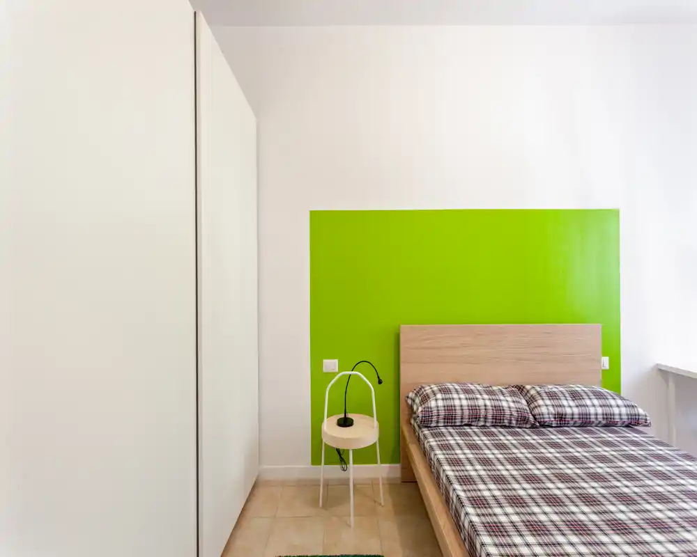 Camera da letto luminosa e minimal con letto in legno e biancheria a quadri su una parete accentuata di verde brillante. Linee pulite, pavimento in piastrella e un piccolo comodino con lampada moderna completano lo spazio ordinato.