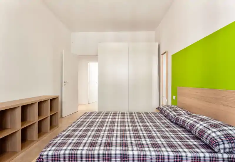 Dormitorio moderno y luminoso con cama doble amplia con ropa de cuadros, cabecero de madera y estantería baja; armario blanco y pared verde vivo que aportan un aspecto fresco y minimalista.