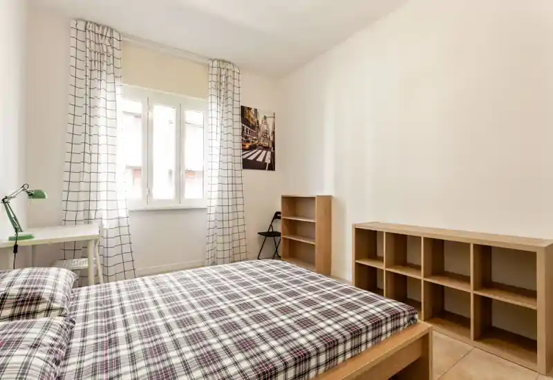 Camera da letto luminosa e arredata con semplicità: letto matrimoniale con copriletto a quadri, finestra con tende a fantasia, scrivania e scaffalature modulari — ideale per un soggiorno accogliente e soleggiato.