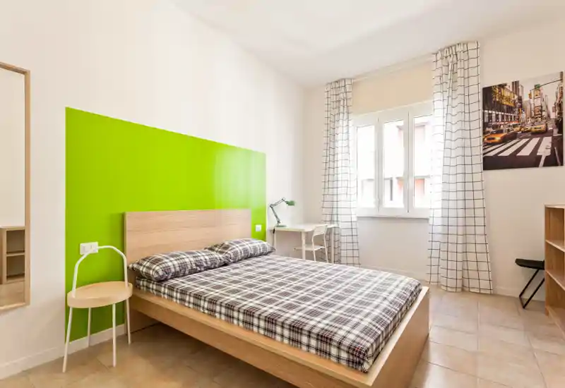 Camera da letto luminosa e moderna con letto matrimoniale, biancheria a quadri e una parete verde vivace; luce naturale dalla finestra e una scrivania semplice, ideale per brevi soggiorni.