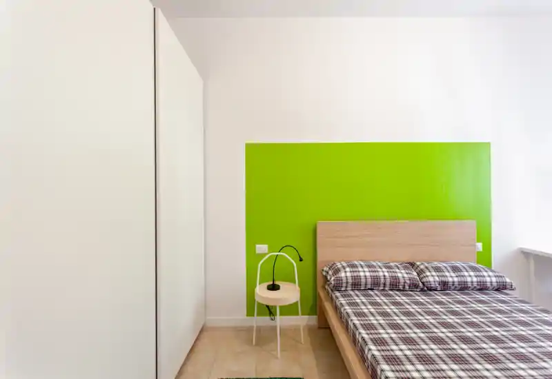 Camera da letto moderna e luminosa con letto in legno e biancheria a quadri davanti a un pannello verde acceso; arredo minimal con un piccolo comodino rotondo e una lampada.
