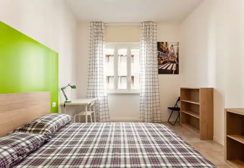 Camera da letto luminosa e ordinata con un ampio letto in primo piano, copriletto a quadri, una scrivania vicino alla finestra e semplici scaffali in legno — ariosa e moderna.