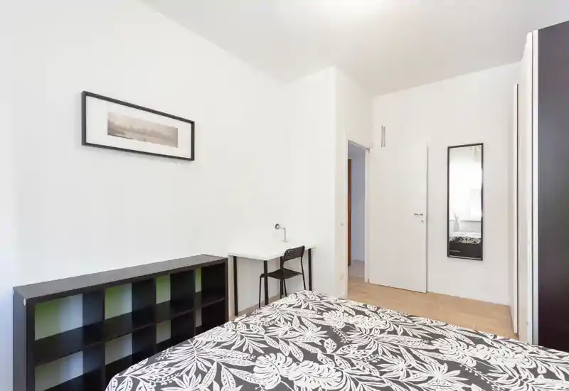 Camera da letto luminosa e minimale con copriletto stampato, una piccola scrivania e scaffale: linee pulite e molta luce naturale, perfetta per annunci.