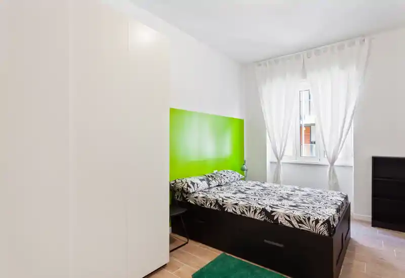 Camera da letto luminosa e moderna con letto contenitore nero, biancheria a fantasia e una vivace testata verde. La luce naturale entra dalle tende leggere, creando un ambiente pulito e arioso perfetto per annunci di affitto.