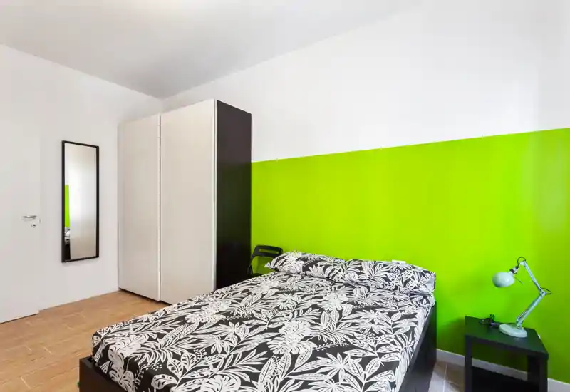 Camera da letto moderna e luminosa con letto matrimoniale dalla biancheria pattern, parete verde a contrasto, armadio e comodino con lampada — stile urbano semplice e fresco.