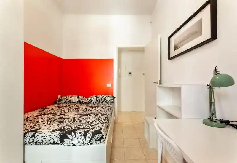 Camera compatta con letto matrimoniale appoggiato a una parete rossa a contrasto, biancheria stampata, scrivania bianca con scaffali e una lampada verde: luminosa, pulita e ben arredata per affitti brevi.
