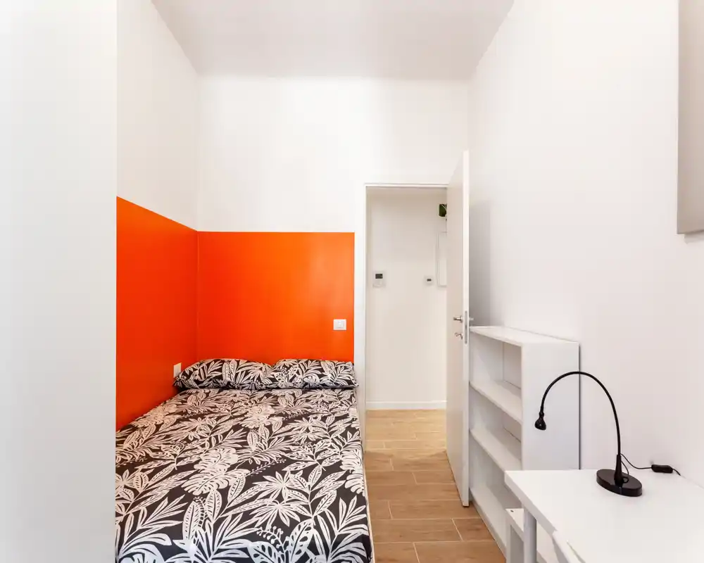 Dormitorio compacto y bien presentado con cama doble frente a una pared de acento naranja, ropa de cama estampada, pequeño escritorio con lámpara y estantería abierta — luminoso y moderno.