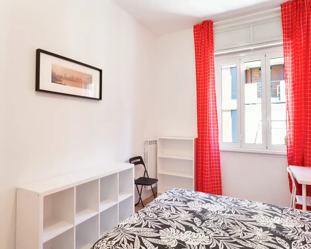 Dormitorio luminoso y sencillo con una cama doble con funda estampada, estanterías blancas y cortinas rojas que enmarcan una ventana grande con abundante luz natural.