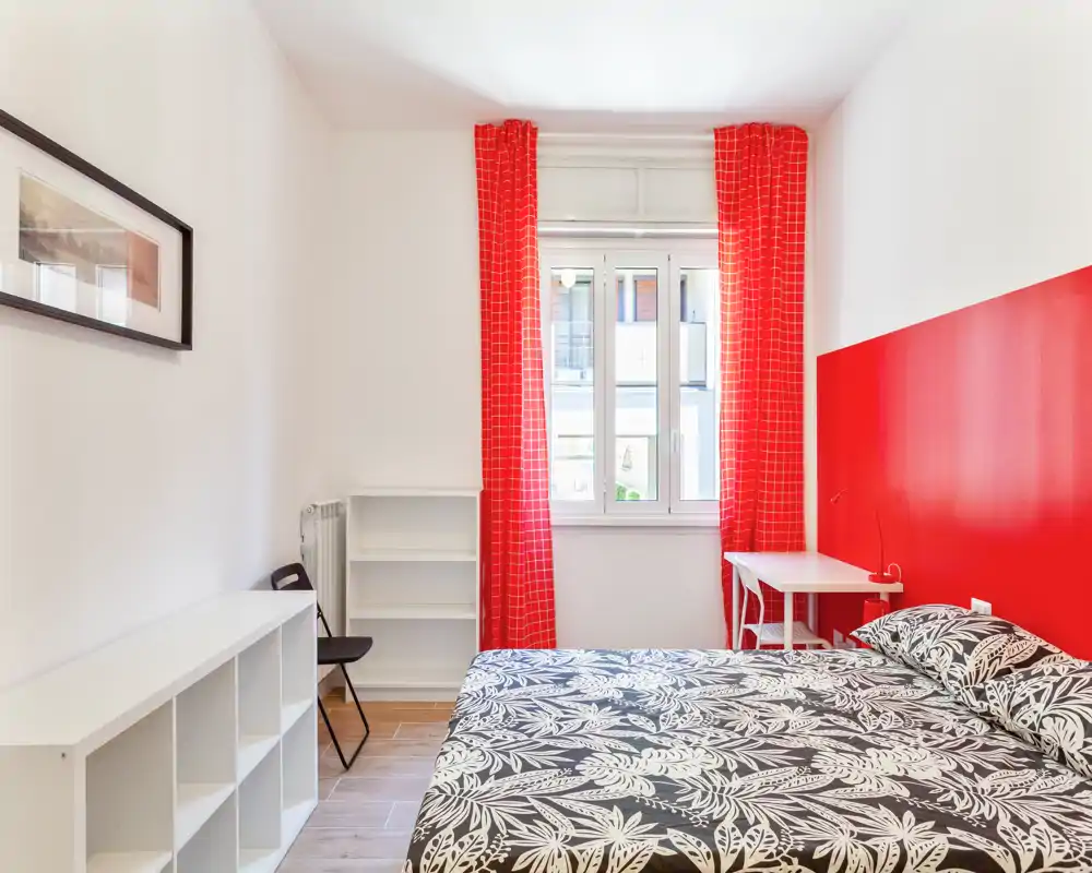 Dormitorio luminoso con cama doble, ropa de cama con estampado y pared y cortinas rojas que destacan. Estanterías sencillas y un pequeño escritorio para almacenamiento y trabajo.