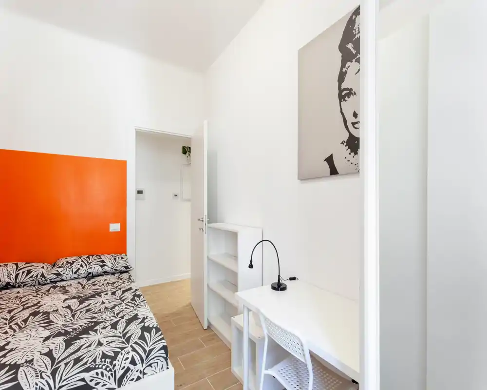 Dormitorio moderno y luminoso con cama doble estampada, pared de acento naranja y escritorio blanco compacto; líneas limpias y buena luz natural, ideal para alquileres de corta estancia.