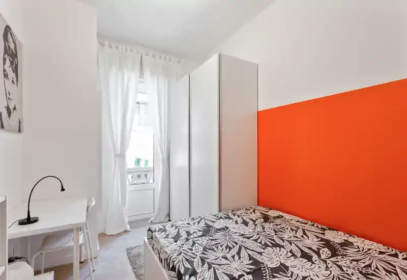 Dormitorio luminoso y compacto con cama doble con ropa de cama estampada, armario blanco y pequeño escritorio junto a la ventana; la pared naranja aporta carácter y buena luz natural.