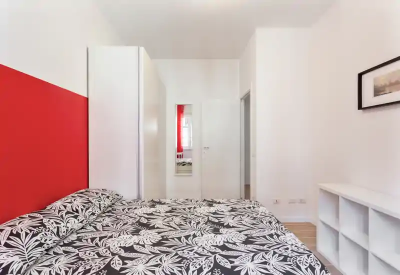 Dormitorio moderno y luminoso con cama doble estampada, pared roja de acento y muebles blancos minimalistas — ordenado y bien iluminado para anuncios.