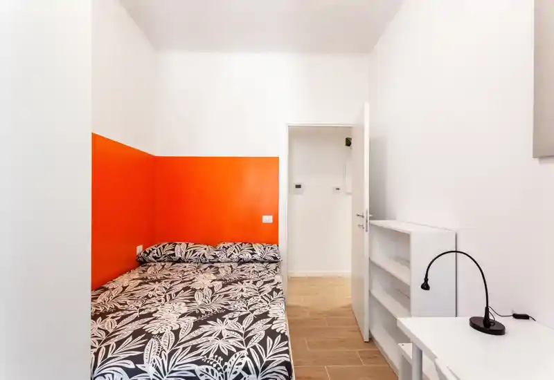 Camera da letto luminosa e compatta con parete d'accento arancione, letto matrimoniale con copriletto a fantasia, scaffalatura semplice e una piccola lampada da scrivania—ideale per affitti brevi moderni.