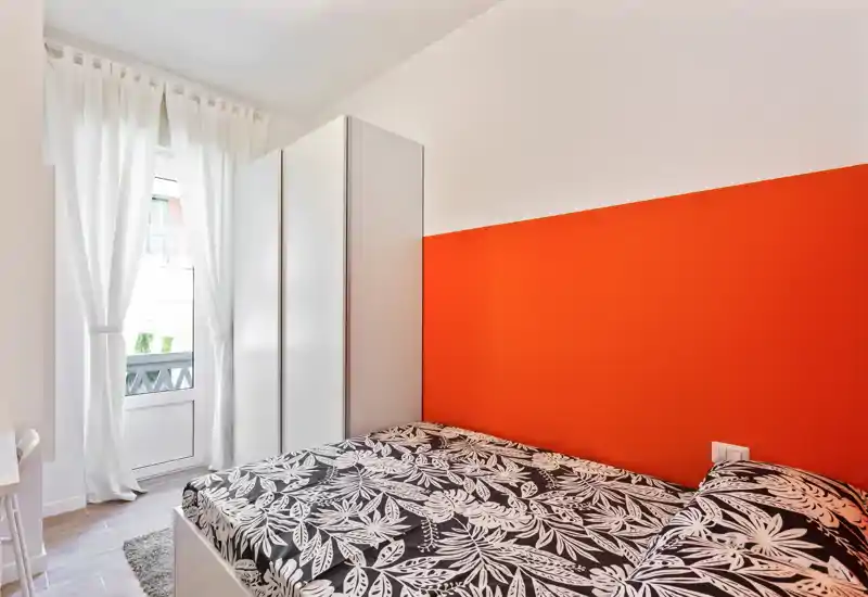 Dormitorio compacto y luminoso con cama doble estampada, pared de acento naranja y un amplio armario junto a la ventana que deja entrar luz natural.