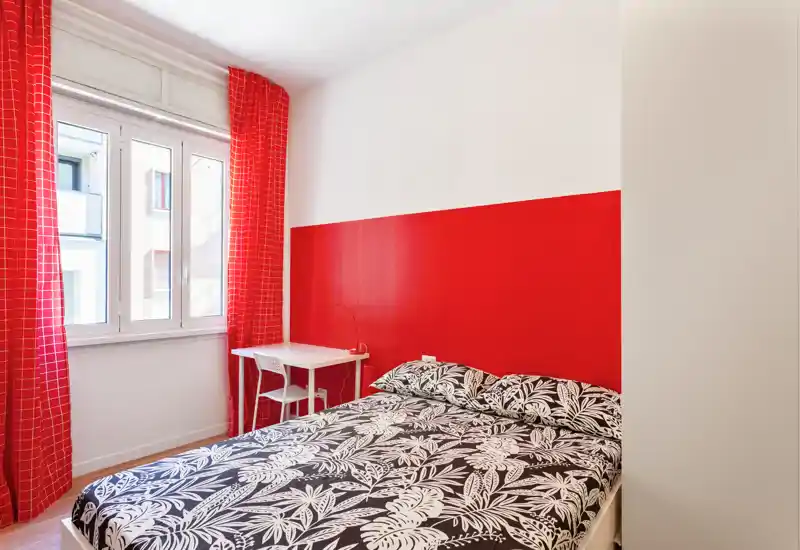 Dormitorio luminoso y acogedor con cama doble frente a una pared roja y cortinas a juego; pequeño escritorio junto a la ventana con buena luz natural.