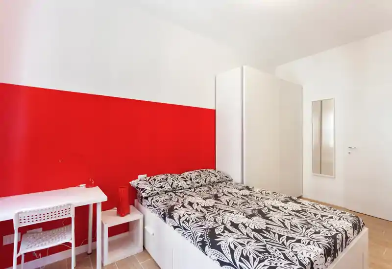 Dormitorio moderno y luminoso con una pared roja de acento, ropa de cama estampada y un pequeño escritorio. Líneas limpias, armario y mesita garantizan un espacio práctico y con estilo para estancias cortas o largas.