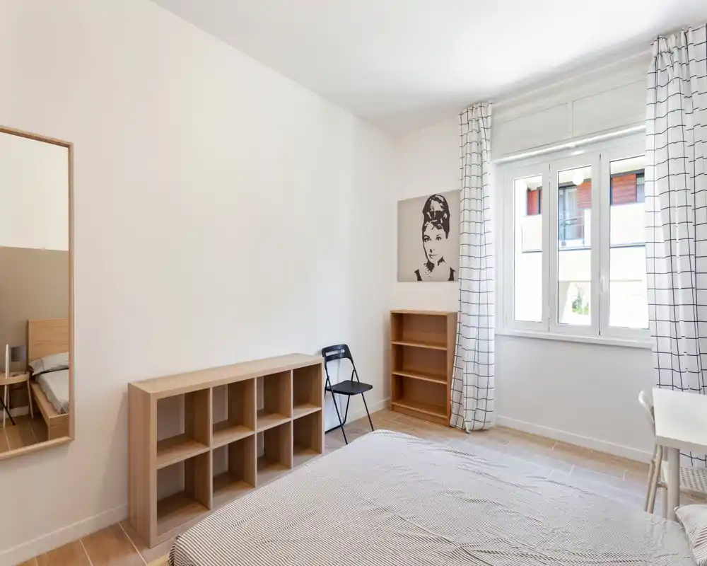 Camera da letto luminosa e minimale con letto ben rifatto, luce naturale da una finestra ampia, scaffalature semplici e una scrivania — ideale per un annuncio moderno.