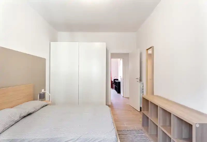 Dormitorio luminoso y minimalista con cama grande, cabecero de madera, armario blanco y puerta abierta hacia el resto del apartamento.