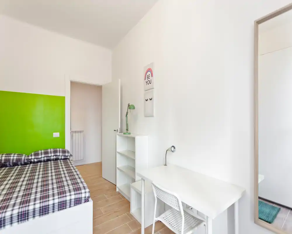 Camera da letto luminosa e ordinata con scrivania bianca e letto davanti a una parete verde; biancheria a quadri, mensole semplici e un grande specchio per un look minimalista.