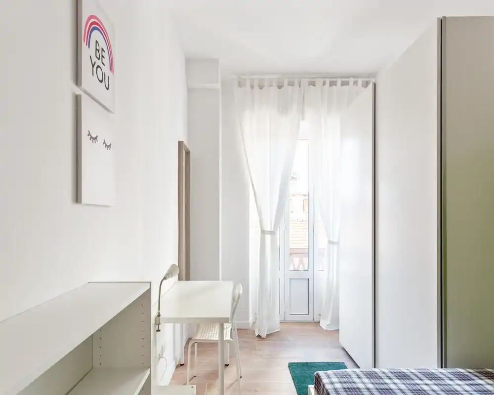 Camera da letto luminosa e minimal con luce naturale, una scrivania vicino alla finestra-balcone e un letto parzialmente visibile con copriletto a quadri: uno spazio pulito e arioso ideale per una persona sola.