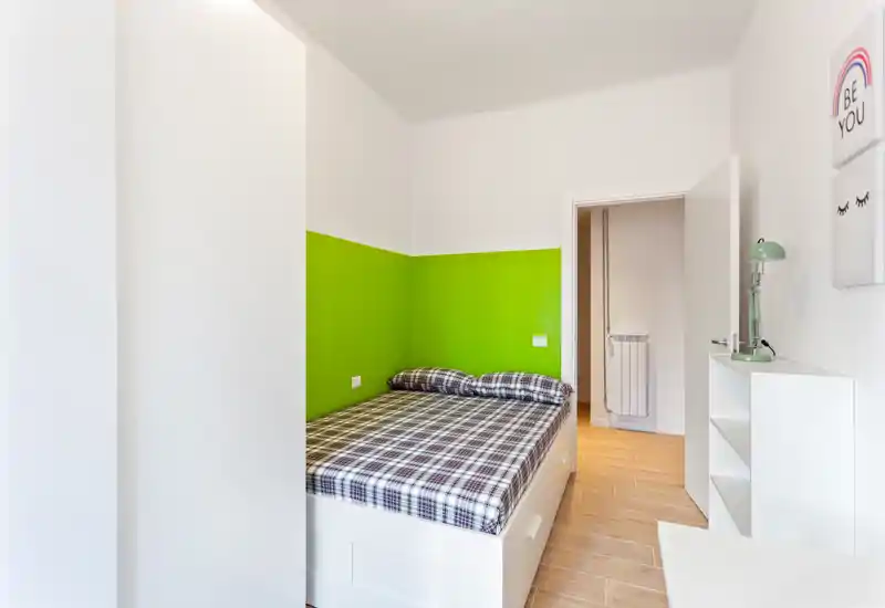 Camera da letto compatta e luminosa con letto matrimoniale davanti a una parete verde acceso; biancheria a quadri, cassetti sotto il letto e una mensola bianca minimal rendono l’ambiente moderno e ordinato.