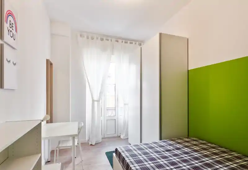 Camera singola luminosa e ordinata con letto coperto da una fantasia a quadri, scrivania bianca e armadio davanti a una finestra alta con tende leggere. Ambiente arioso e minimal con una parete accentata verde.