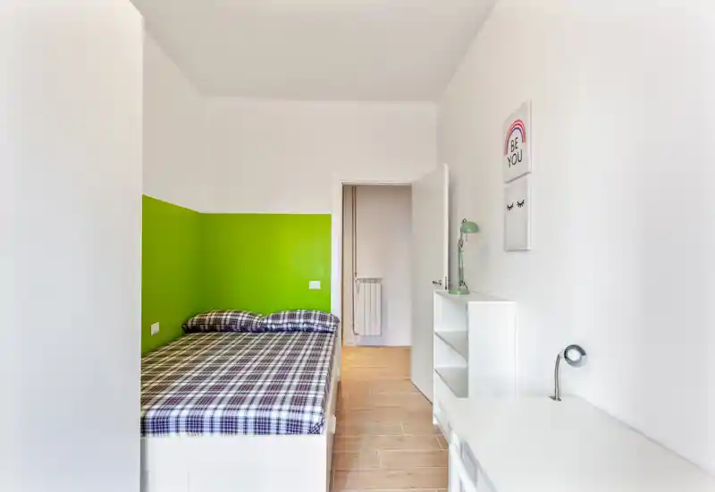 Camera da letto luminosa e compatta con letto matrimoniale a sinistra, parete verde a contrasto e arredi bianchi essenziali tra cui scrivania e scaffale. Stile pulito, moderno e ben illuminato, ideale per una persona o una coppia.
