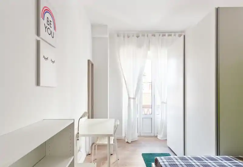 Camera da letto luminosa e minimal con luce naturale, una scrivania vicino alla finestra-balcone e un letto parzialmente visibile con copriletto a quadri: uno spazio pulito e arioso ideale per una persona sola.