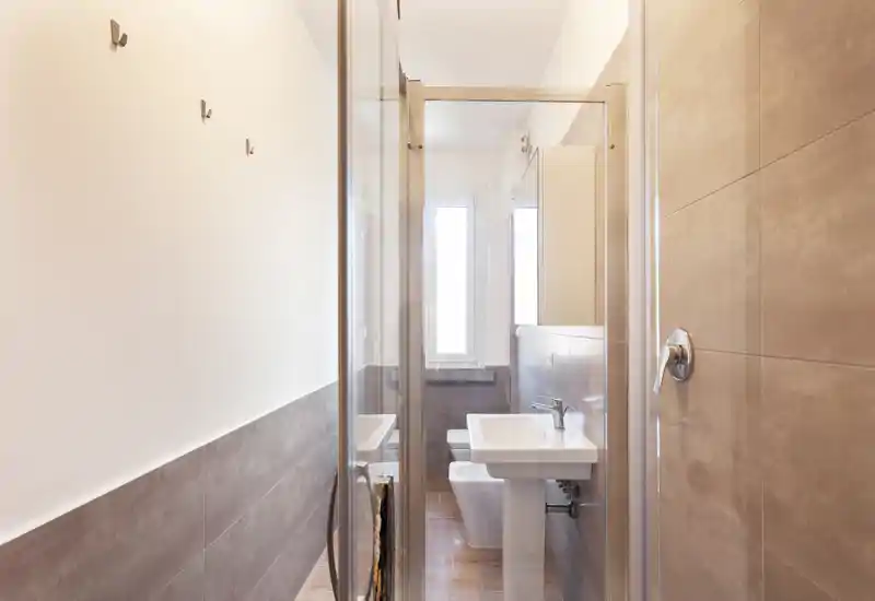 Baño moderno y luminoso con lavabo de pie, inodoro y puerta de ducha de cristal; azulejos neutros y luz natural de una ventana alta.