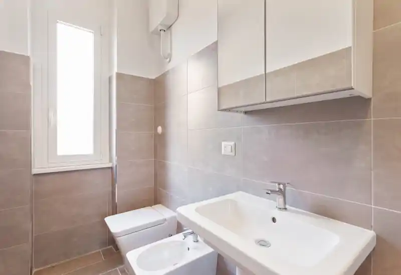 Baño moderno y luminoso con mueble de espejo, lavabo, bidé y WC. Azulejos neutros y una ventana alta esmerilada que aporta luz natural suave.