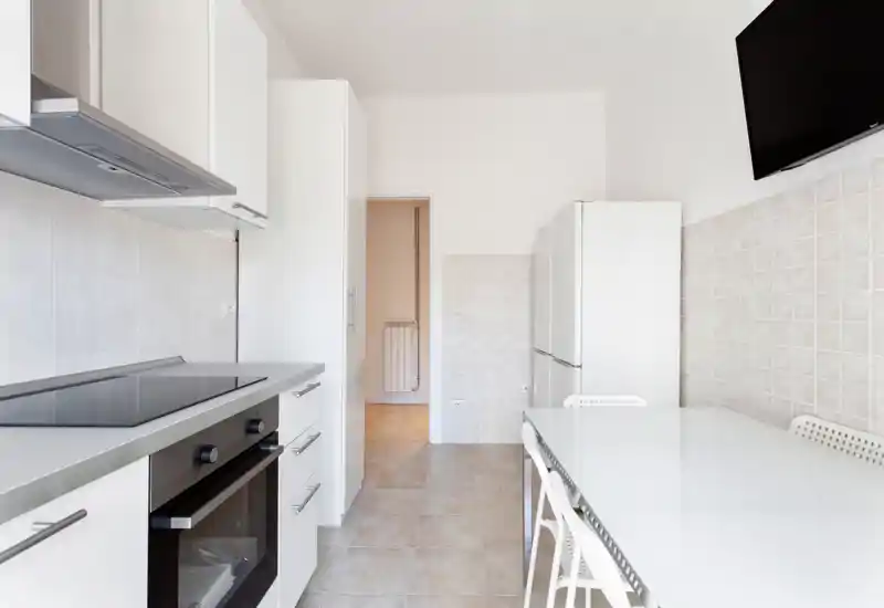 Cocina moderna y luminosa tipo pasillo con horno integrado, placa de inducción y frigorífico alto. Muebles blancos y una mesa sencilla crean un espacio de cocina práctico y minimalista.