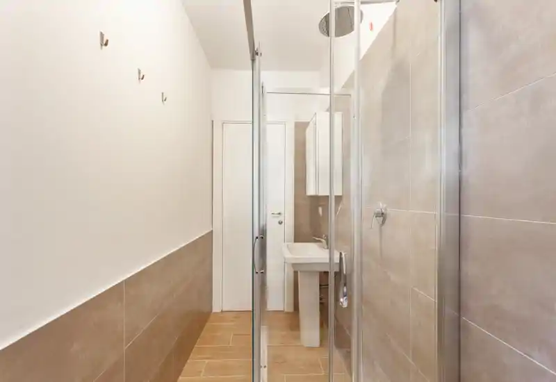 Baño moderno y compacto con mampara de ducha de cristal, lavabo de pedestal y azulejos beige neutro. Espacio luminoso, limpio y bien iluminado, adecuado para un anuncio de alquiler.