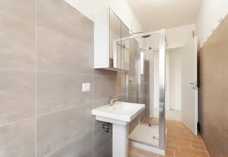 Baño moderno y luminoso con lavabo de pedestal, armario con espejo y ducha acristalada; azulejos neutros y suelo efecto madera para un aspecto limpio y contemporáneo.