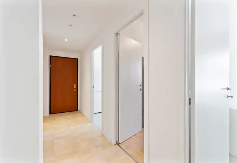 Vestíbulo de entrada luminoso y minimalista con puerta principal de madera y suelo de baldosas, paredes blancas neutras y varias puertas interiores hacia las habitaciones adyacentes.